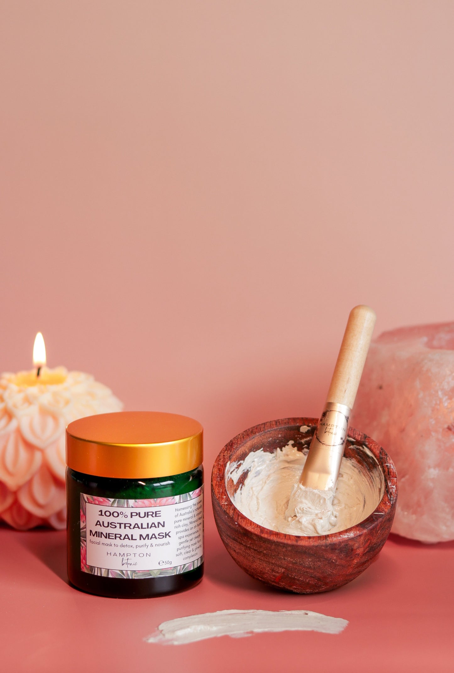 A peach coloured dahlia shaped candle, a jar of clay mask next to a wooden bowl with clay mask in it and a mask brush, and a pink Himalayan salt candle holder behind it, all on a peach coloured background